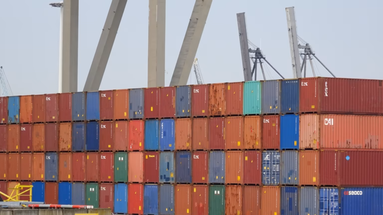 Standard shipping containers stacked four high at the Port of Rotterdam, with crane booms rising behind them