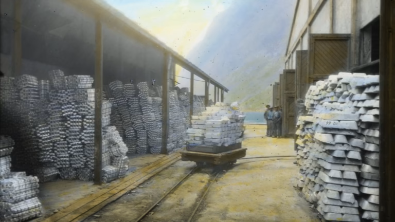 Pile di lingotti di alluminio e un carrello su binari nel magazzino della Norsk Aluminium Company a Høyanger, Norvegia, in una diapositiva colorata a mano del 1926