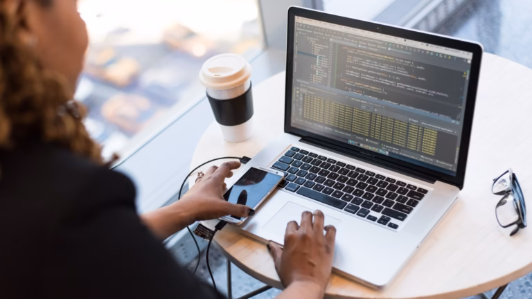 A developer at a laptop reading source code and log output in an IDE, with a phone connected for testing