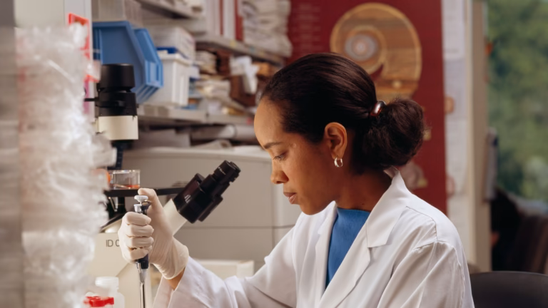 A researcher in a white lab coat and gloves works with a pipette beside a microscope in a biomedical laboratory