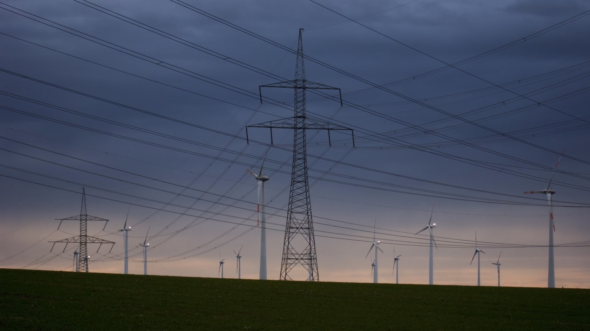High-voltage pylons and overhead lines crossing a wind farm under an overcast evening sky near Nauen, Brandenburg, Germany.