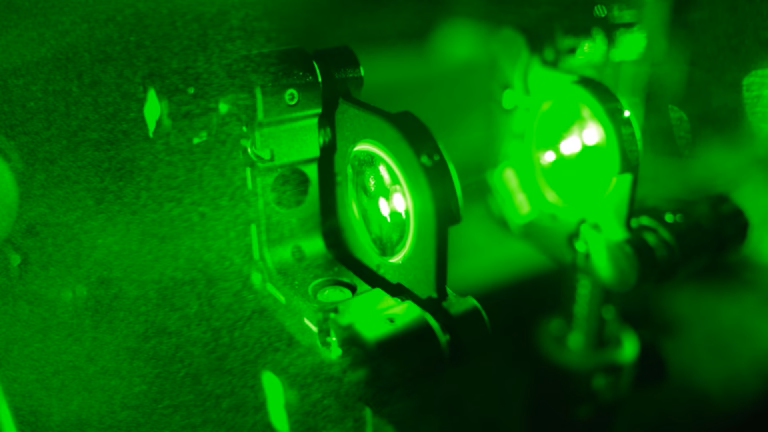 A green laser beam passing through a lens doublet held in mounts on an optical bench, the lenses glowing in a dark laboratory
