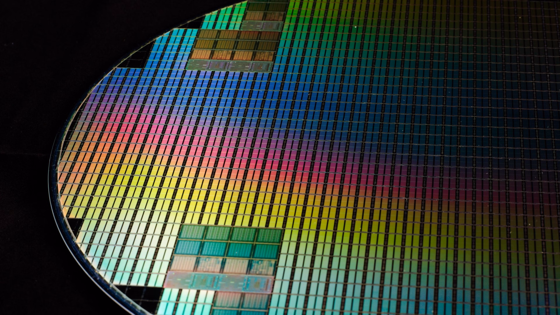 Close-up of a patterned silicon wafer, its grid of chips glowing in iridescent colours against a black background