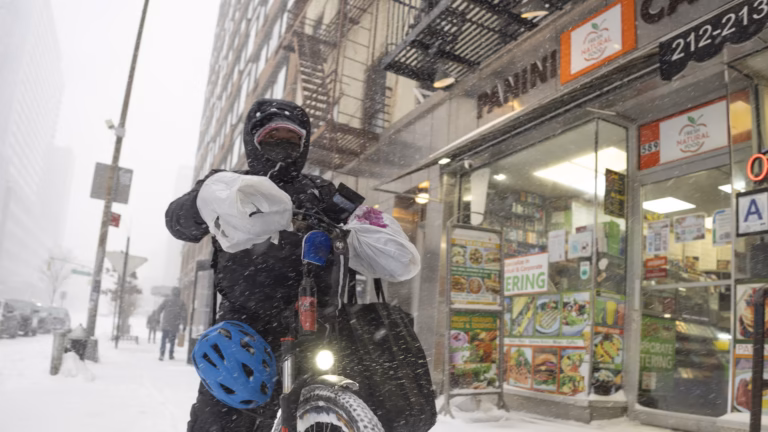 A delivery courier wrapped against the cold walks a bicycle loaded with bags through a snowstorm on a city sidewalk