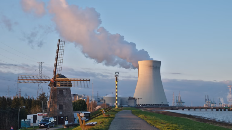 A nuclear power plant cooling tower venting steam at dusk beside a brick windmill and high-voltage pylons at Doel, Belgium