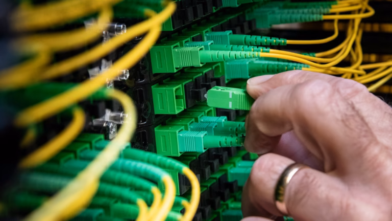 A technician's hand seating a fibre optic jumper into a dense panel of green connectors in a data centre