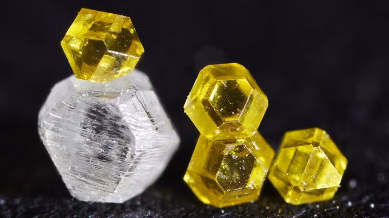 Macro photograph of lab-grown synthetic diamond crystals, one colourless and four yellow from nitrogen impurities, the smallest about 0.3 mm across