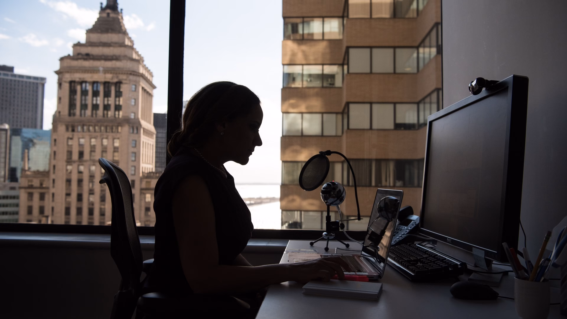 Silhouette of a person working at a laptop and monitor beside a high-rise office window overlooking the city