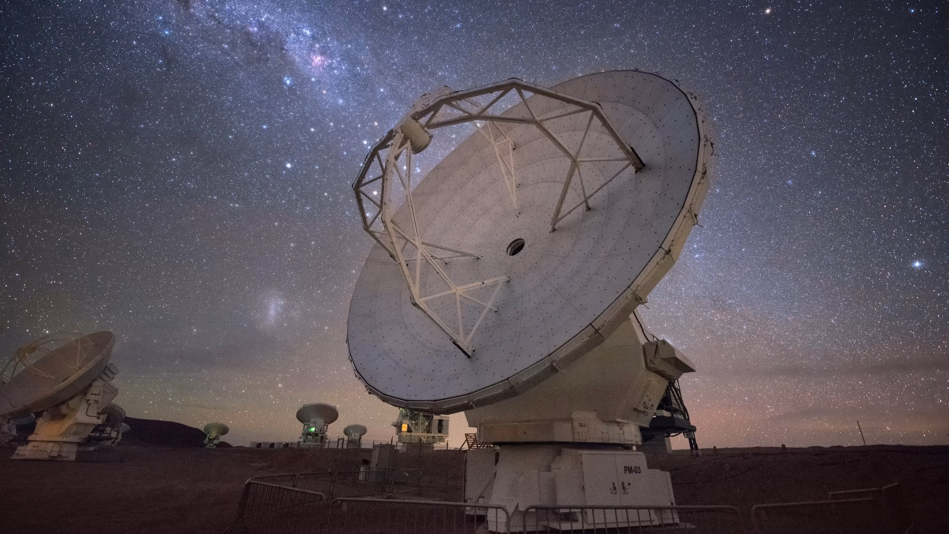 A radio telescope dish of the ALMA observatory in Chile's Atacama Desert, tilted toward a star-filled night sky with the Milky Way and the Magellanic Clouds